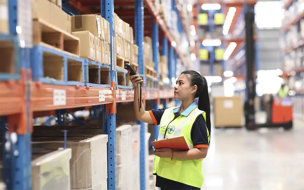 Lady wearing a hi viz and holding a clipboard scanning an item on a shelf in a warehouse